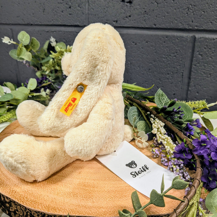 Beige teddy bear with a visible brand tag on a wooden block surrounded by greenery and flowers.