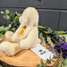 Beige teddy bear with a visible brand tag on a wooden block surrounded by greenery and flowers.