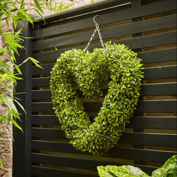 Heart-shaped green wreath hanging on a black slatted wall with foliage around.