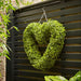 Heart-shaped green wreath hanging on a black slatted wall with foliage around.