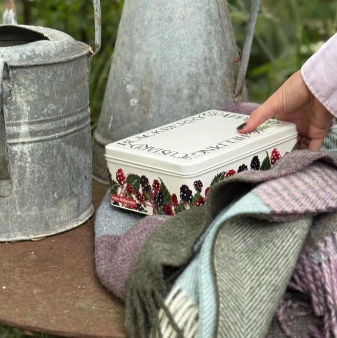 Decorative tin box with blackberry design on a wooden surface with a rustic metal watering can in the background.