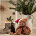 Brown teddy bear wearing a Santa hat with Christmas decorations including pine branches and pinecones.