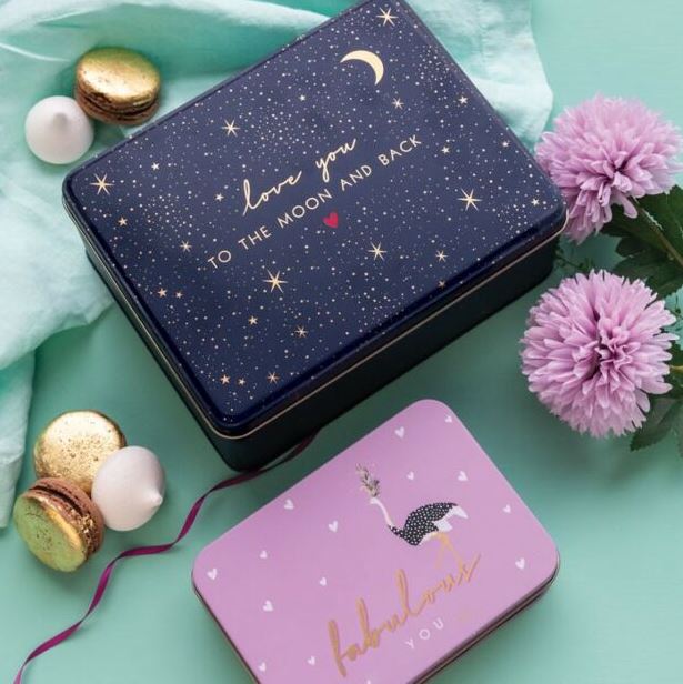 Two decorative tins with romantic phrases on a teal background with flowers and macarons.