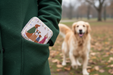 Person wearing a green coat with a decorative treat tin in their pocket, walking a dog in a park.