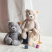 Two teddy bears, one gray and one beige, sitting on a white surface with a soft pink curtain in the background.