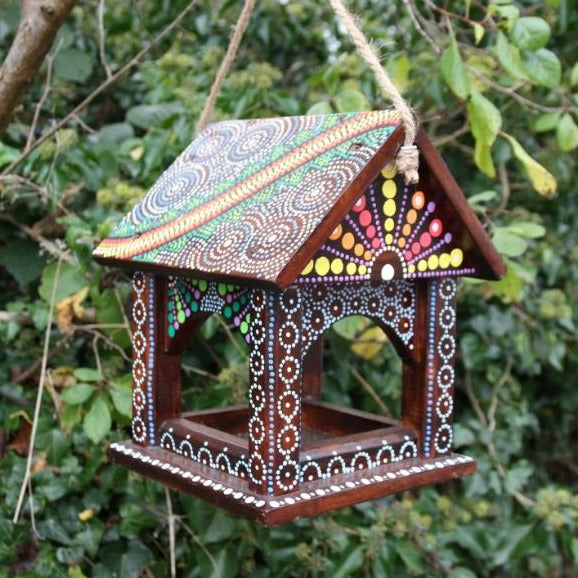 bird feeding table hung from a tree branch, the bird feeder is painted in various dot patterns in bright colours. 