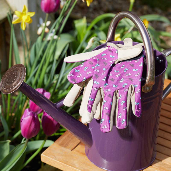 Purple gardening gloves with butterflies on the back resting on a purple watering can next to a flower bed. 