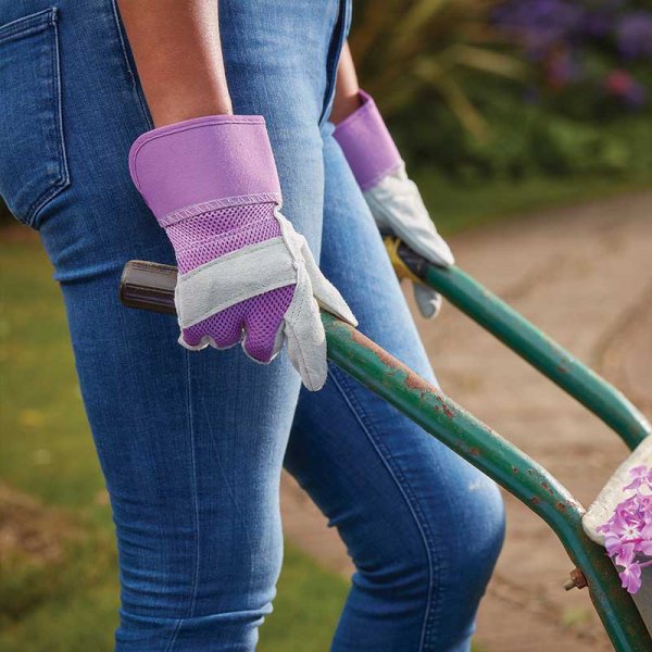 Purple and grey gardening gloves being used by a women pushing a wheel barrow in a garden. 