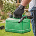 Black and grey gardening gloves being used by a man holding a fuel can in a garden setting. 