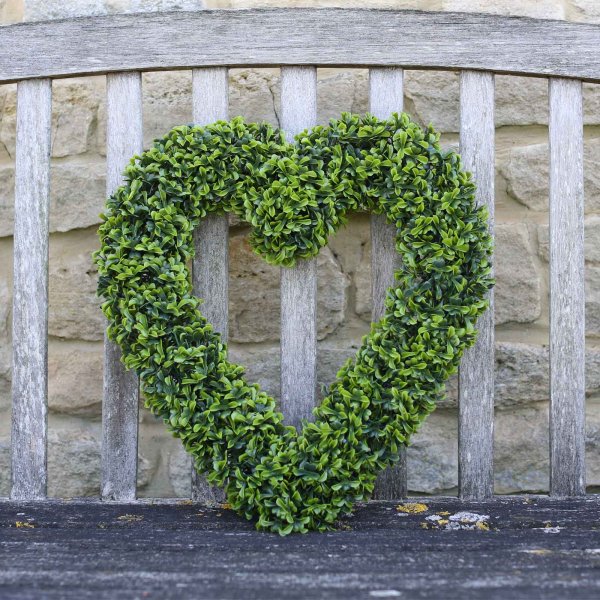 Artificial topiary heart shaped wreath propped against an outdoor wooden bench.